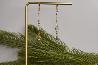 Gold earrings with gemstones on a gold stand with green foliage in the background