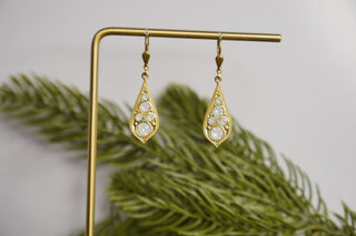 Gold earrings with teardrop design on a gold stand with green leaves in the background