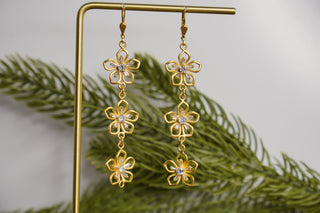 Gold flower-shaped earrings with clear stones on a stand with green leaves in the background