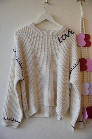 Beige sweater with black text on a hanger against a beige wall