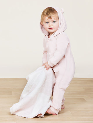 Child wearing a pink onesie with a hood on a light wooden floor.