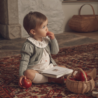 Child sitting on a patterned rug with a book and apples, surrounded by a cozy indoor setting.