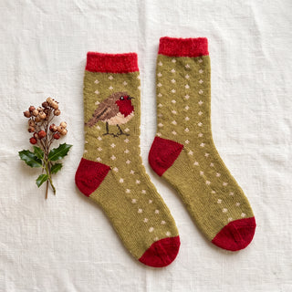 A pair of olive green knit socks with red cuffs, heels, and toes, featuring a small robin bird motif on one sock and a pattern of small white flowers on the other. The socks are displayed on a textured white fabric background next to a small sprig of holly leaves and berries.