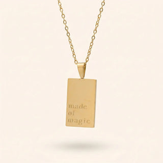 Gold necklace with a rectangular pendant engraved 'made of magic' on a beige background