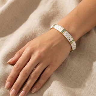 Hand wearing a white bracelet with gold and green accents on a beige fabric background