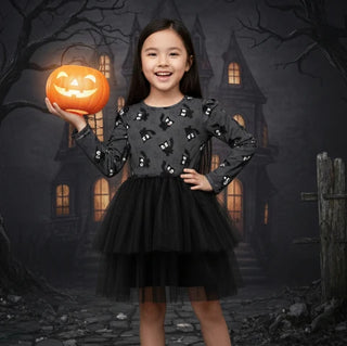 A young girl wearing a black long-sleeve Halloween dress with a tulle skirt stands in front of a spooky haunted-house backdrop, holding an orange jack-o'-lantern.