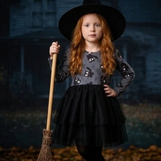 A young girl with long red hair wearing a black witch hat and a black and gray long-sleeve dress with bat patterns, holding a broomstick in front of a spooky house background.