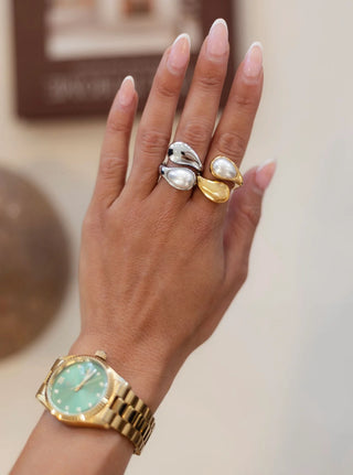 Hand wearing a gold watch and two pearl rings on a neutral background