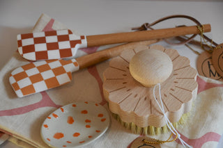 Set of kitchen utensils including checkered spatulas, a wooden brush, and a polka dot dish on a patterned cloth.