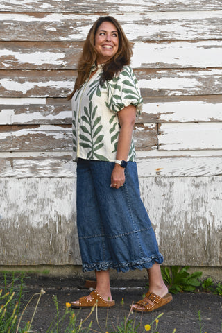A woman stands outdoors in front of a weathered white wooden wall, wearing a cream short-sleeve blouse with a green leaf print, a blue denim skirt, and brown studded slide sandals.