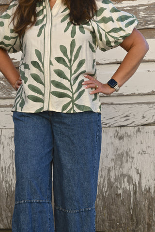 A woman wearing a cream-colored short-sleeve blouse with a green leaf pattern and blue wide-leg jeans, standing in front of a weathered white wooden wall.