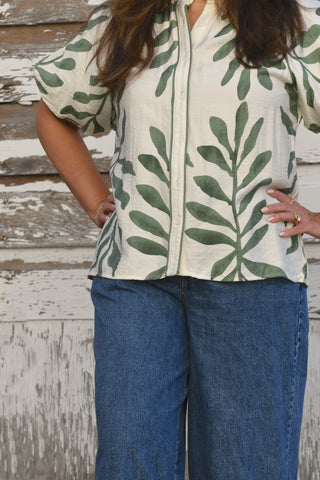 A woman wearing a cream-colored short-sleeve button-up shirt with a green leaf pattern, paired with blue jeans, standing against a rustic white-painted wooden wall.