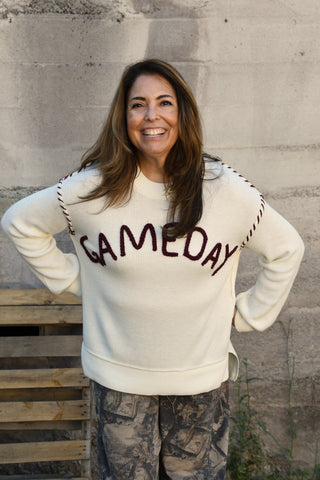 A woman smiling and posing outdoors in front of a concrete wall, wearing a cream-colored crewneck sweatshirt with 'GAMEDAY' written across the chest in dark brown lettering, paired with camouflage pants.