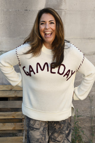 A woman smiling and laughing while wearing a cream-colored knit sweater with 'GAMEDAY' written across the chest in dark brown lettering, paired with camouflage pants, standing against a concrete wall.