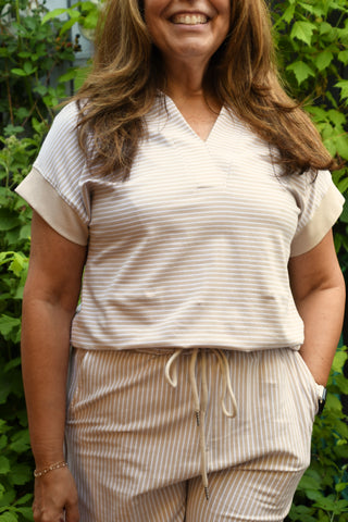 A woman wearing a matching beige and white striped pajama set, featuring a short-sleeve V-neck top and drawstring pants, standing outdoors in front of green foliage.