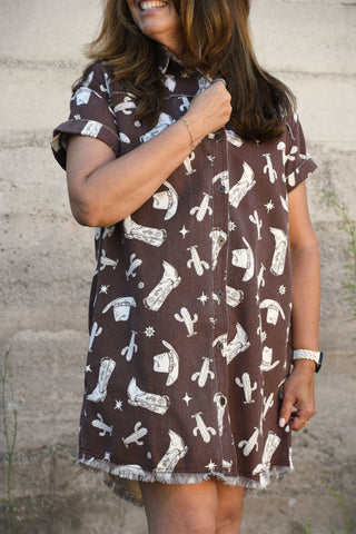 A woman wearing a brown short-sleeve button-up shirt dress with a white western-themed print of boots, hats, and cacti, standing against a textured concrete wall.