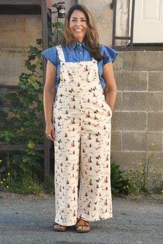 A woman wearing a cream wide-leg jumpsuit with a brown and red botanical print, styled over a blue short-sleeve button-up shirt, standing outdoors in front of a stone wall.