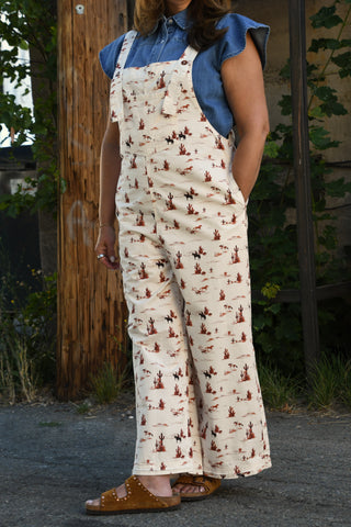 A person wearing cream wide-leg bib overalls with a brown cowboy print, styled over a blue denim shirt and brown slide sandals, standing outdoors next to a wooden post.