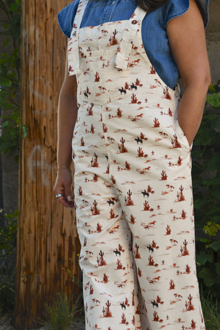 A person wearing cream-colored bib overalls with a brown and black western desert print, layered over a blue denim top, standing outdoors next to a wooden post.