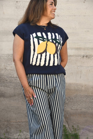 Woman wearing a navy sweater with lemon design and striped pants against a concrete wall.