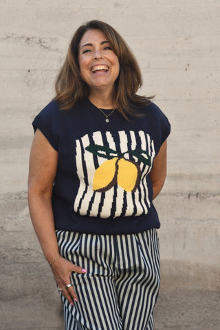 Woman wearing a navy sweater with lemon design and striped pants against a beige wall.