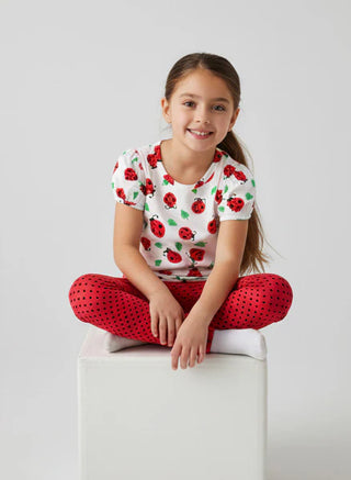 Young girl sitting on a white stool wearing a ladybug-patterned shirt and red polka dot pants.