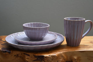 A matching ceramic tableware set in a muted lavender-purple color, featuring a mug with a ribbed handle, a small bowl, and two plates with a matching saucer, arranged on a rustic wood slab against a plain gray wall.