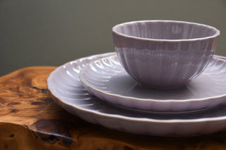 A matching ceramic dinnerware set with a small ribbed bowl, two stacked plates, and a matching saucer, all in a muted lavender-gray glaze, displayed on a rustic wooden surface against a soft gray background.