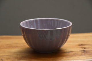 A small lavender-purple ceramic bowl with a ribbed, scalloped exterior, sitting on a wooden tabletop against a neutral gray background.