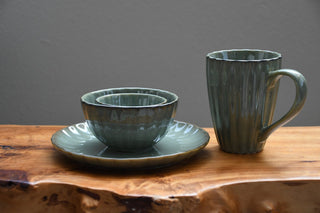A set of handmade ceramic tableware in a muted sage green glaze, including a mug, a matching saucer, and a nested bowl, displayed on a rustic live-edge wooden table against a solid gray background.