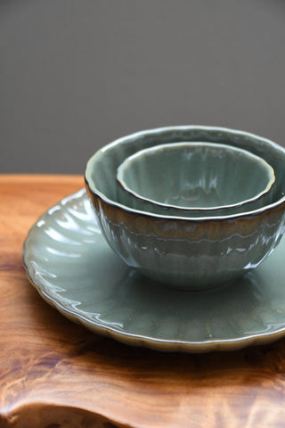 A set of nested ceramic bowls and a matching plate in a muted sage green glaze, resting on a rustic wooden surface against a dark gray background.