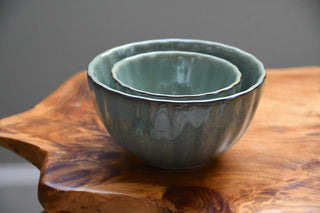 Two nested ceramic bowls with a glossy sage green and gray glaze, displayed on a rustic wooden slab against a neutral gray background.