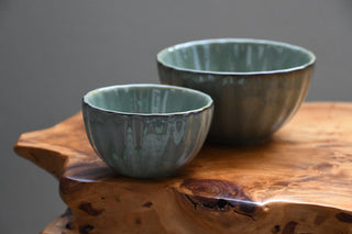 Two handmade ceramic bowls with a glossy, textured sage green glaze, displayed on a rustic live-edge wooden slab against a neutral gray background.