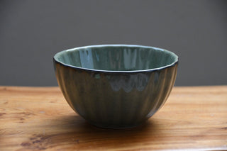 A handmade ceramic bowl with a textured, ribbed exterior and a glossy, speckled teal-green interior, resting on a wooden surface against a dark gray background.