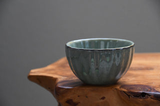 A handmade ceramic bowl with a textured, ribbed exterior and a glossy, mottled sage green glaze, resting on a rustic live-edge wooden table against a neutral gray background.