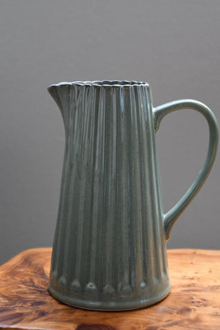 A ribbed ceramic pitcher with a muted sage green glaze, resting on a wooden surface against a solid gray background.