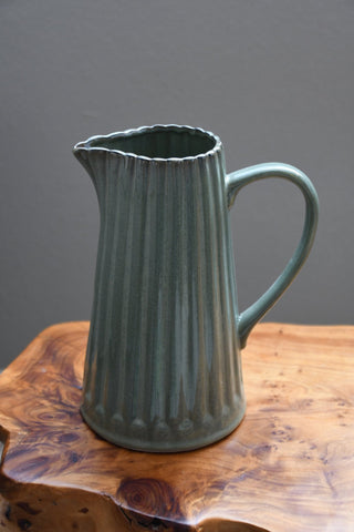 A ribbed ceramic pitcher with a muted sage green glaze, resting on a rustic live-edge wooden table against a solid gray background.