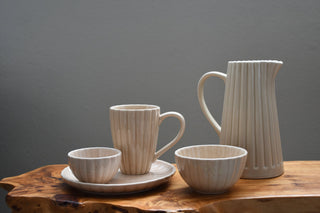 A ceramic tableware set with a ribbed texture, including a pitcher, a mug on a saucer, and two bowls, arranged on a rustic wood slab against a dark gray background.