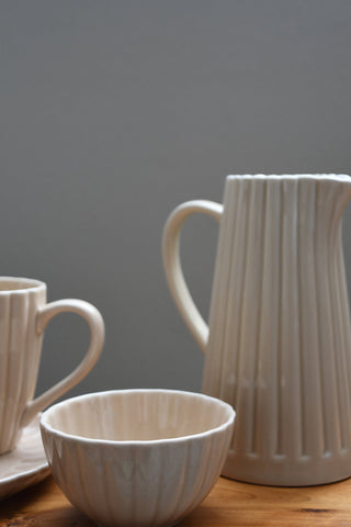 A ceramic tableware set with a ribbed pitcher, bowl, and cup, shown on a wooden surface against a neutral gray background.