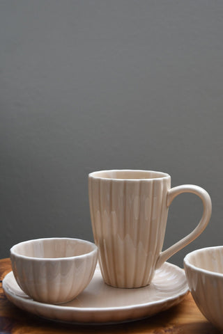 A ceramic mug with a matching saucer and two small bowls, all in a light beige color with a vertical ribbed texture, arranged on a wooden surface against a dark gray background.