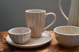A ceramic tableware set in a warm cream-beige ribbed glaze, featuring a mug on a saucer, a small bowl, and another bowl, arranged on a wooden surface against a dark gray background.