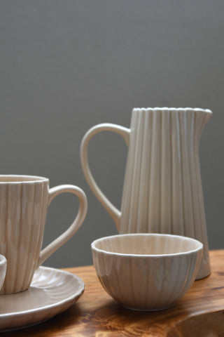 A ceramic tableware set with a ribbed texture in a warm off-white or cream color, including a pitcher, a bowl, a cup on a saucer, and a small dish, arranged on a wooden surface against a neutral gray background.