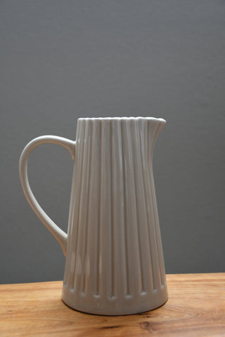 A ribbed ceramic pitcher in a light warm gray or off-white glaze, featuring a curved handle and a spout, resting on a wooden surface against a solid gray background.