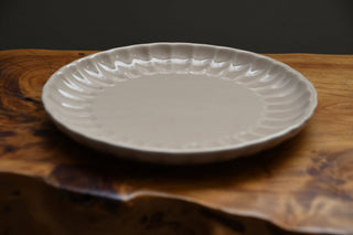 A round off-white ceramic plate with a scalloped rim, displayed on a rustic wooden surface against a dark gray background.