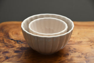 A set of three nested ceramic bowls with a fluted, ribbed exterior and smooth, matte off-white interiors, displayed on a rustic wooden surface against a dark gray background.