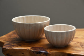 Two ribbed ceramic bowls in a warm off-white glaze, one large and one small, displayed on a rustic live-edge wooden board against a neutral gray background.