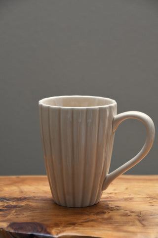 A light beige ceramic mug with a fluted vertical rib pattern and a C-shaped handle, resting on a rustic wooden surface against a solid dark gray background.