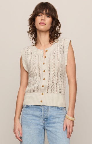 Woman wearing a beige sleeveless knitted top with buttons and light blue jeans on a plain background