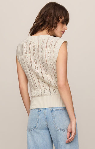 Woman wearing a beige sleeveless knitted top and light blue jeans on a plain background