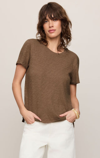 Woman wearing a brown t-shirt and white pants against a plain background